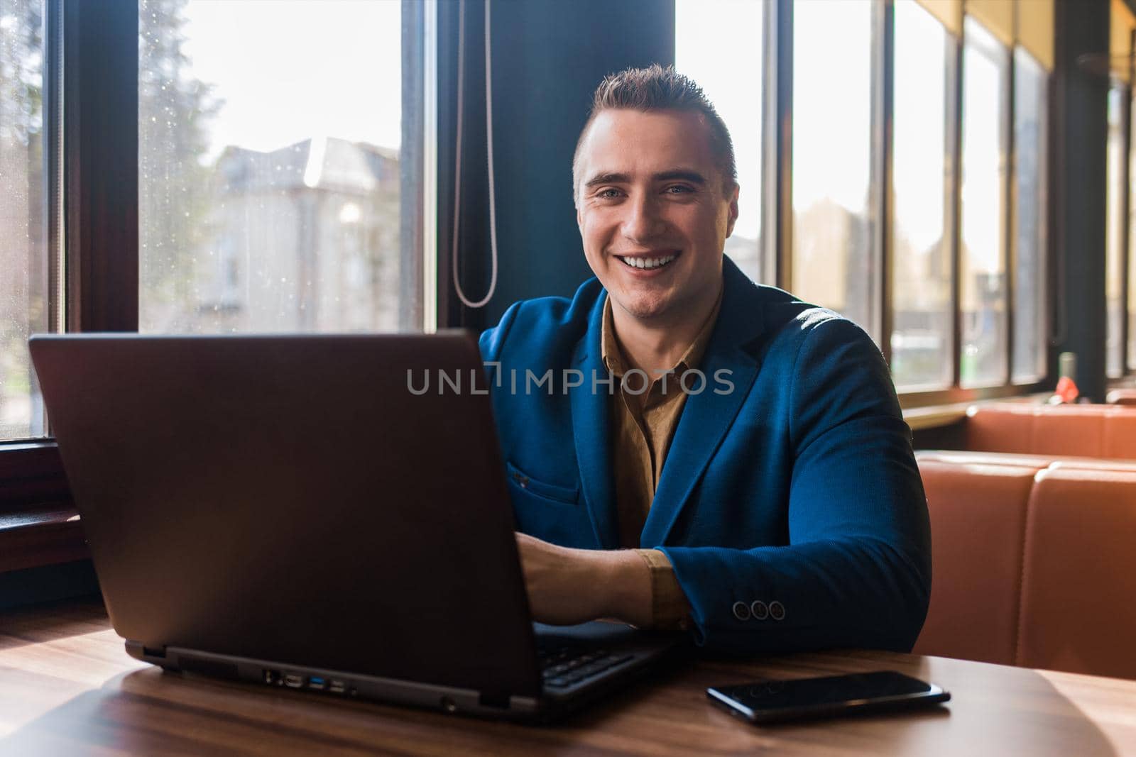 A business smiling man happy positive businessman a stylish portrait of Caucasian appearance in a jacket works in a laptop or computer, sitting at a table by the window in a cafe.