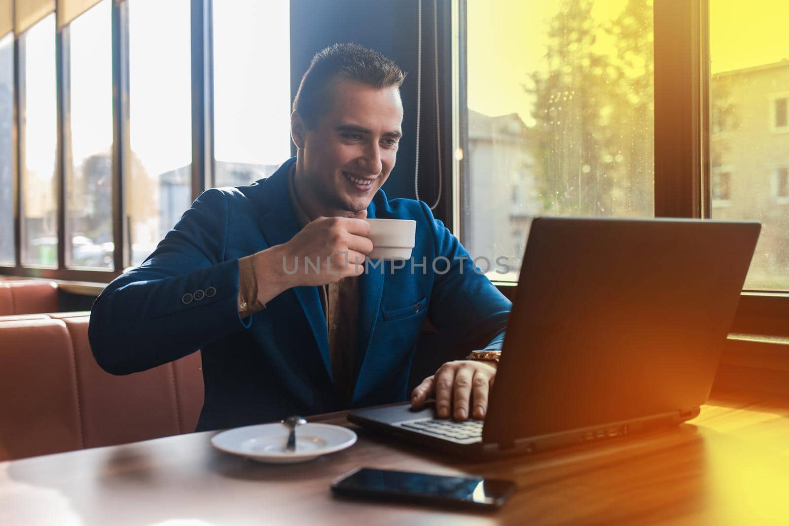 A business smiling positive man businessman a stylish portrait of Caucasian appearance in a jacket, works in a laptop or computer, drink coffee on coffee break, sitting at a table by the window.