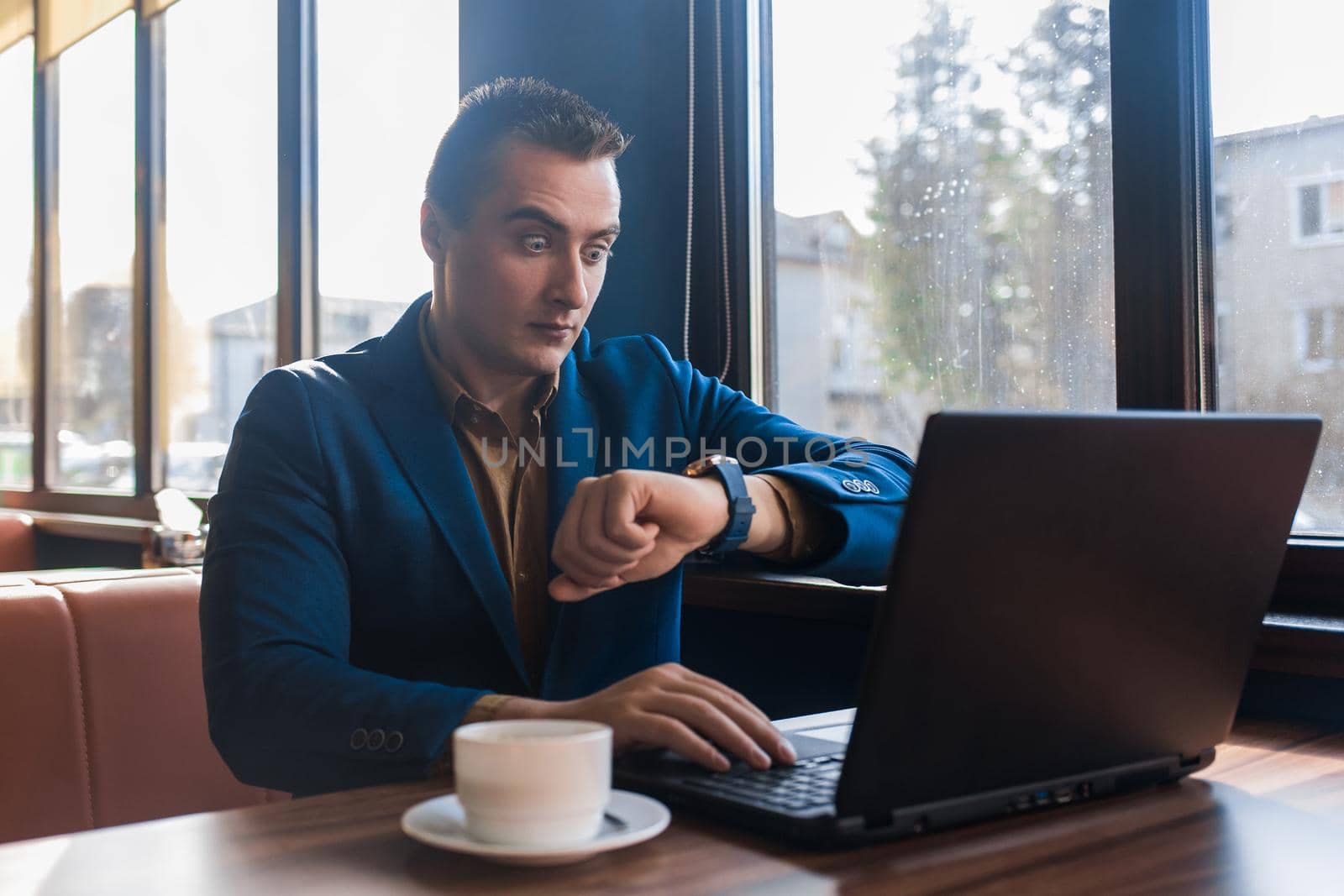 A business surprised man businessman a stylish of Caucasian appearance in a jacket, works in a laptop or computer, sitting at a table by the window in a cafe and looks at the time on a wrist watch.