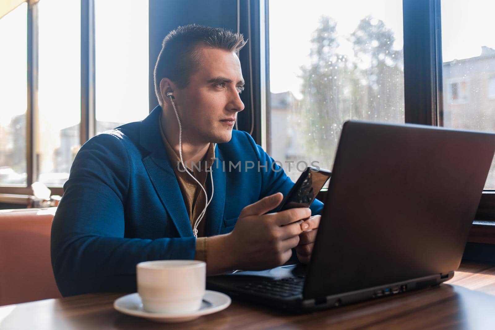 A business pensive man stylish businessman in an attractive European-looking suit works in a laptop, listens to music with headphones and sitting at a table in a cafe by the window.