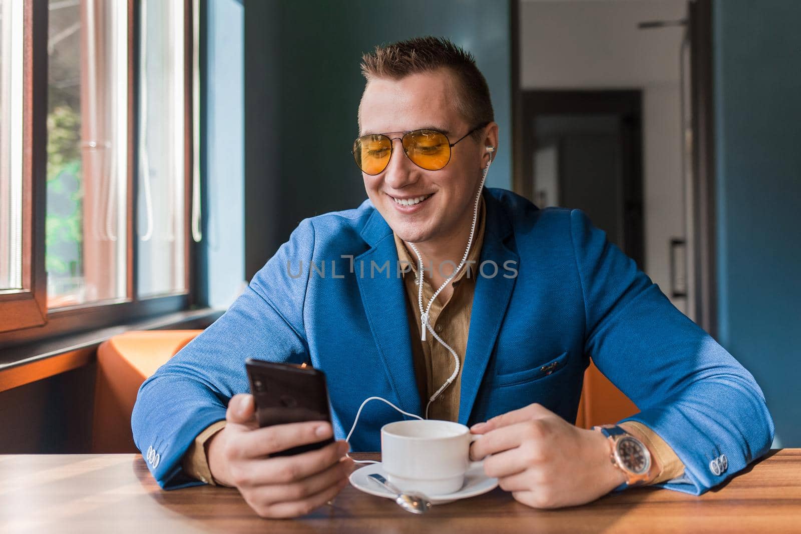 A businessman a stylish, smiling, happy portrait of Caucasian appearance in sunglasses, suit, sits at a table on a coffee break in smartphone and headphones, listen to music on cellphone or mobile in a cafe.