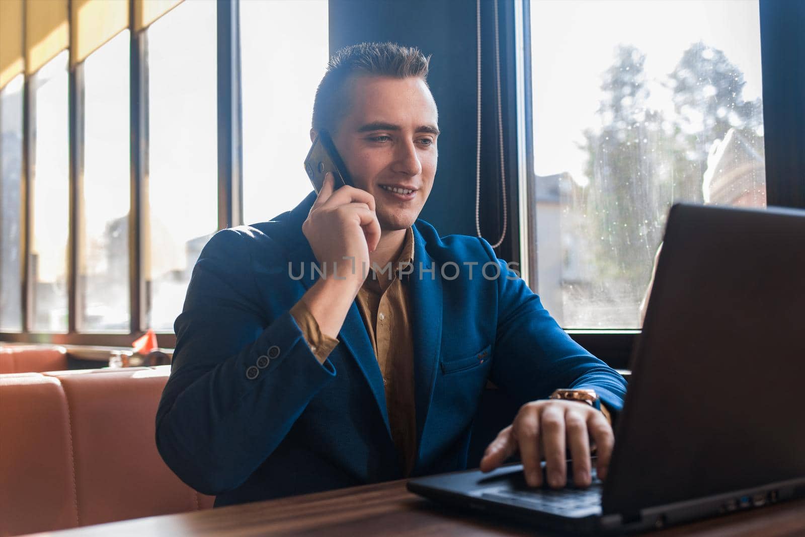 A business man stylish positive smiling businessman in an attractive European-looking suit works in a laptop, talks on a cell or mobile phone, sitting at a table in a cafe by the window.