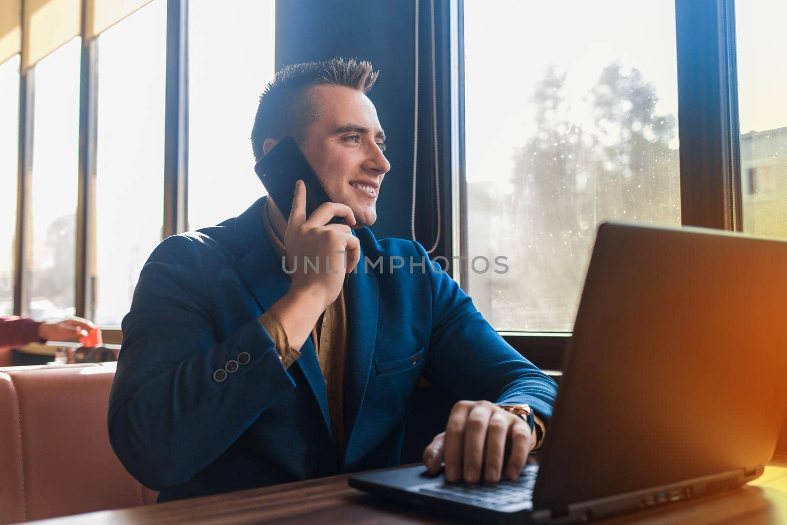 A business man stylish positive smiling businessman in an attractive European-looking suit works in a laptop, talks on a cell or mobile phone, sitting at a table in a cafe by the window.