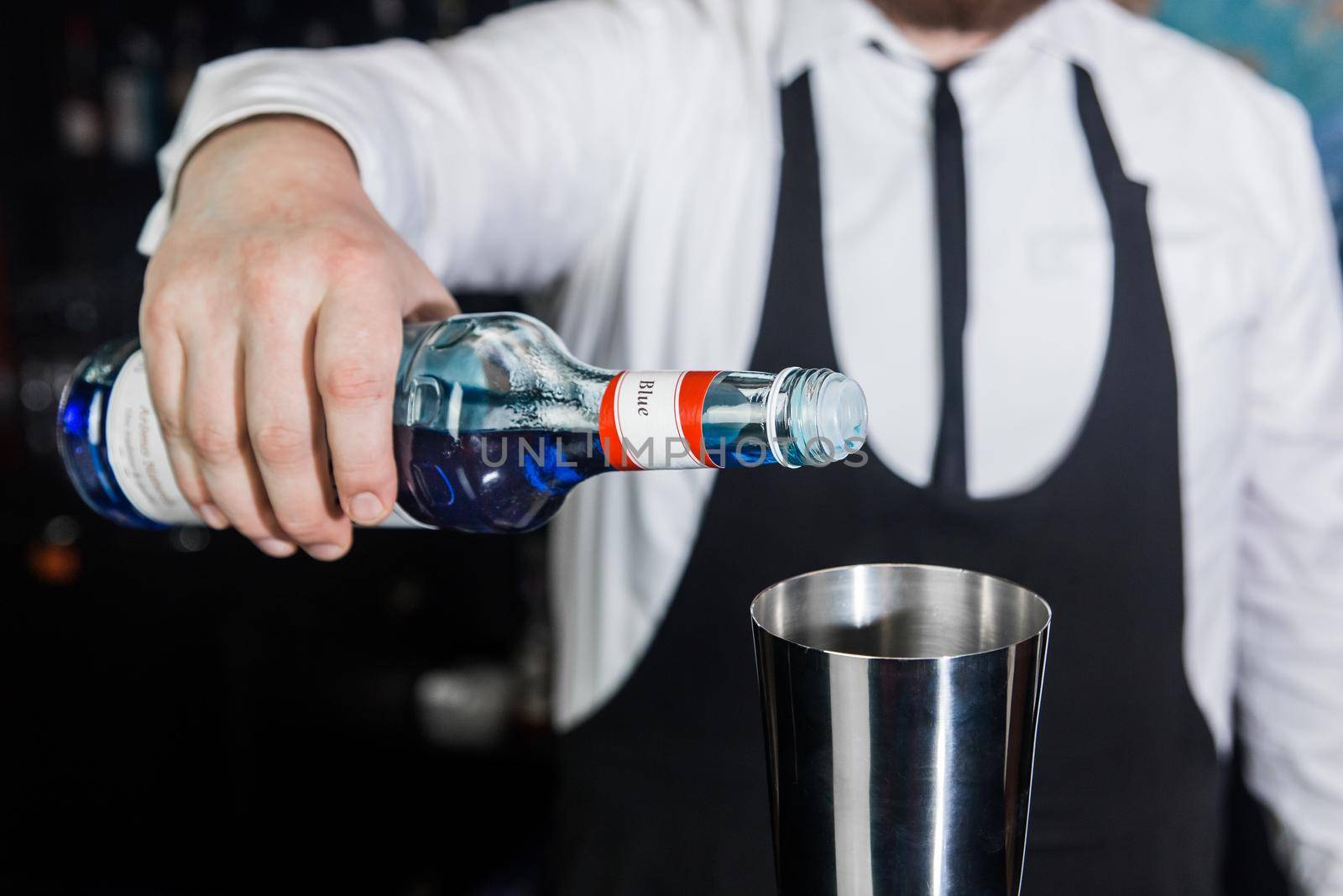 The hand of a professional bartender pours blue syrup into a tool for preparing and mixing alcoholic cocktails shaker, close-up.