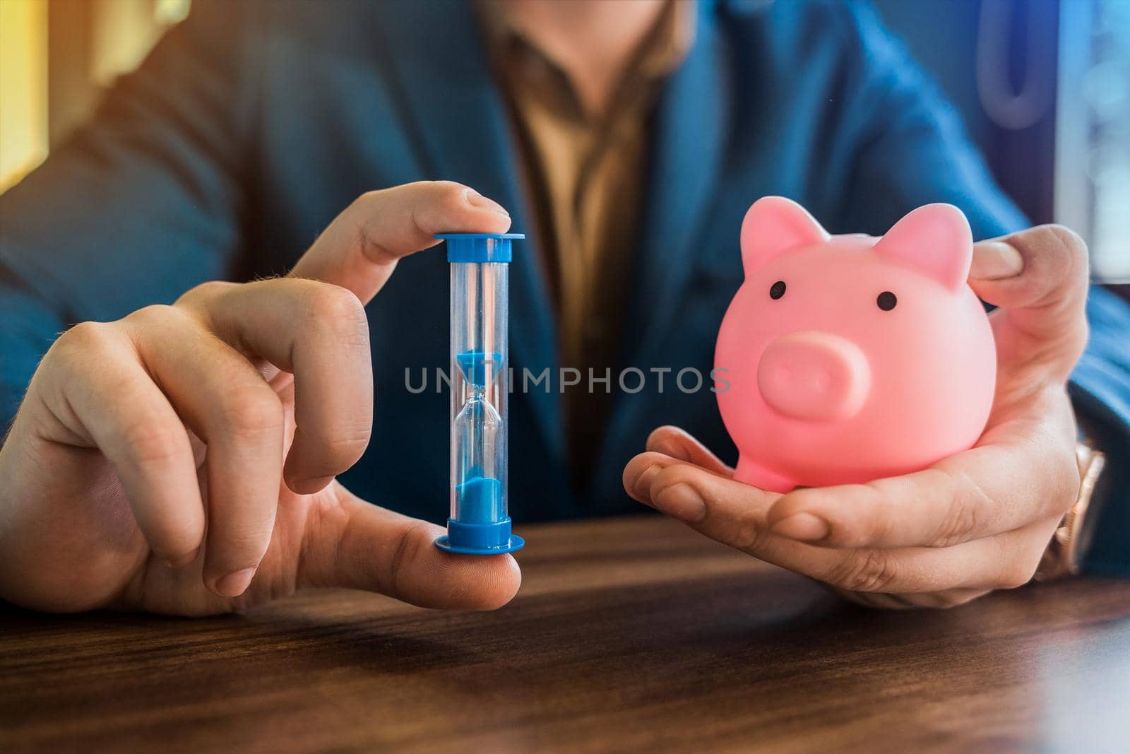 The hands of a smiling businessman in a suit hold an hourglass of time and a piggy bank of savings.