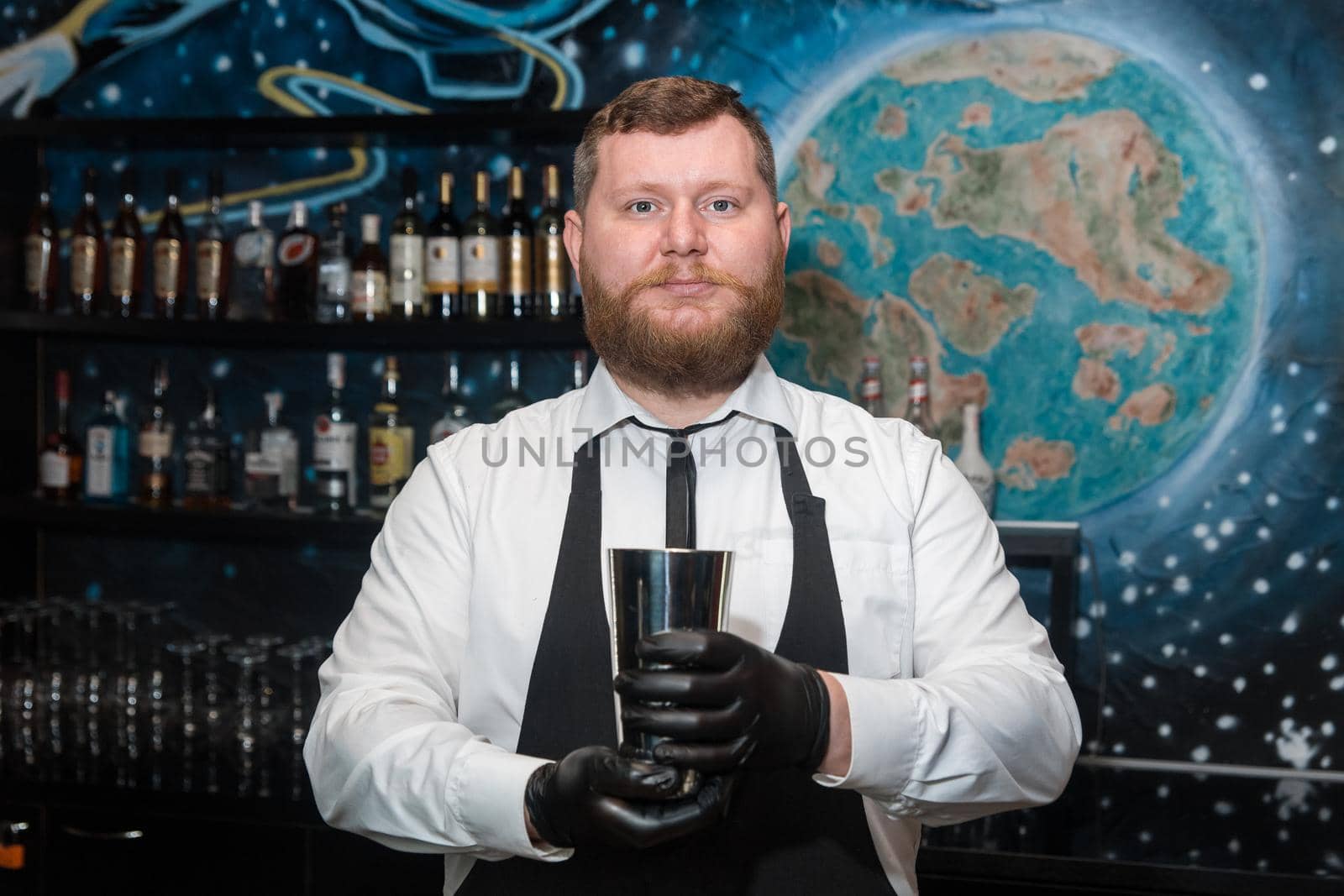 A bearded adult man of European appearance, a professional bartender, holds in his hands in black latex gloves a tool for preparing and mixing alcoholic cocktails in a nightclub.