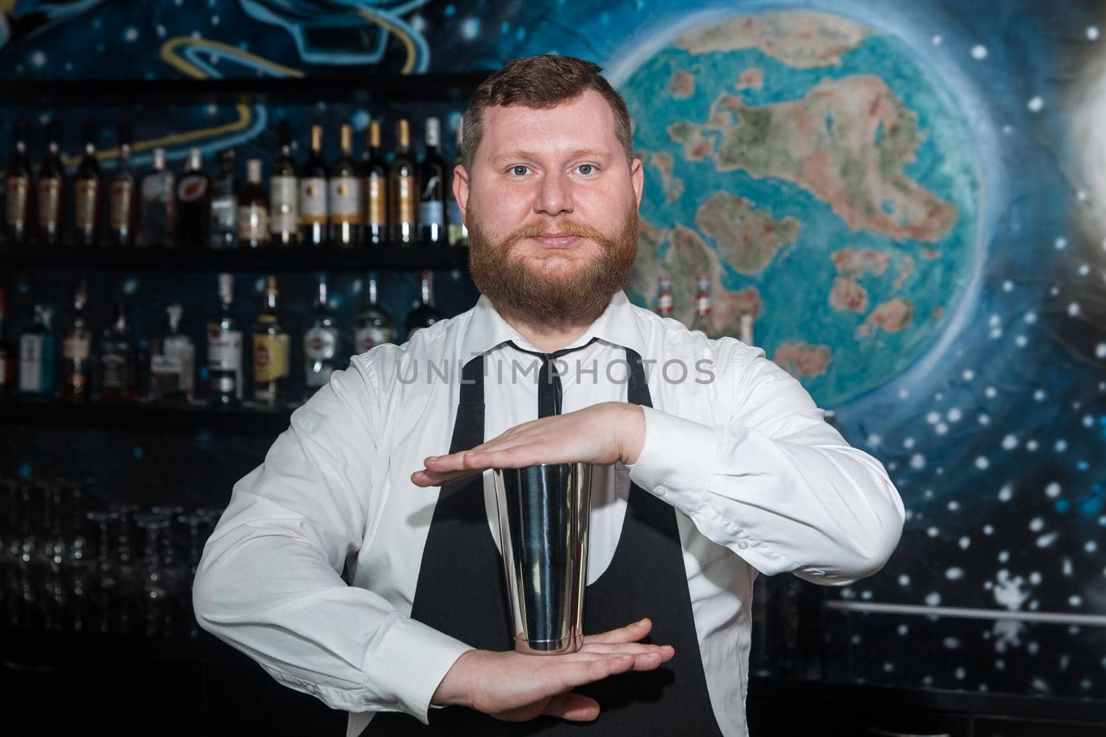 A bearded adult man of European appearance, a professional bartender, holds in his hands a tool for preparing and mixing alcoholic cocktails in a nightclub.