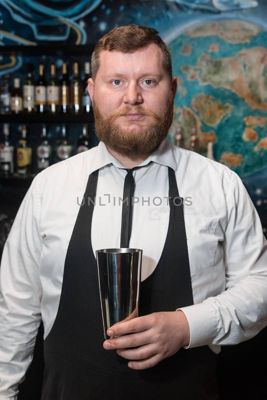 A bearded adult man of European appearance, a professional bartender, holds in his hands a tool for preparing and mixing alcoholic cocktails in a nightclub.