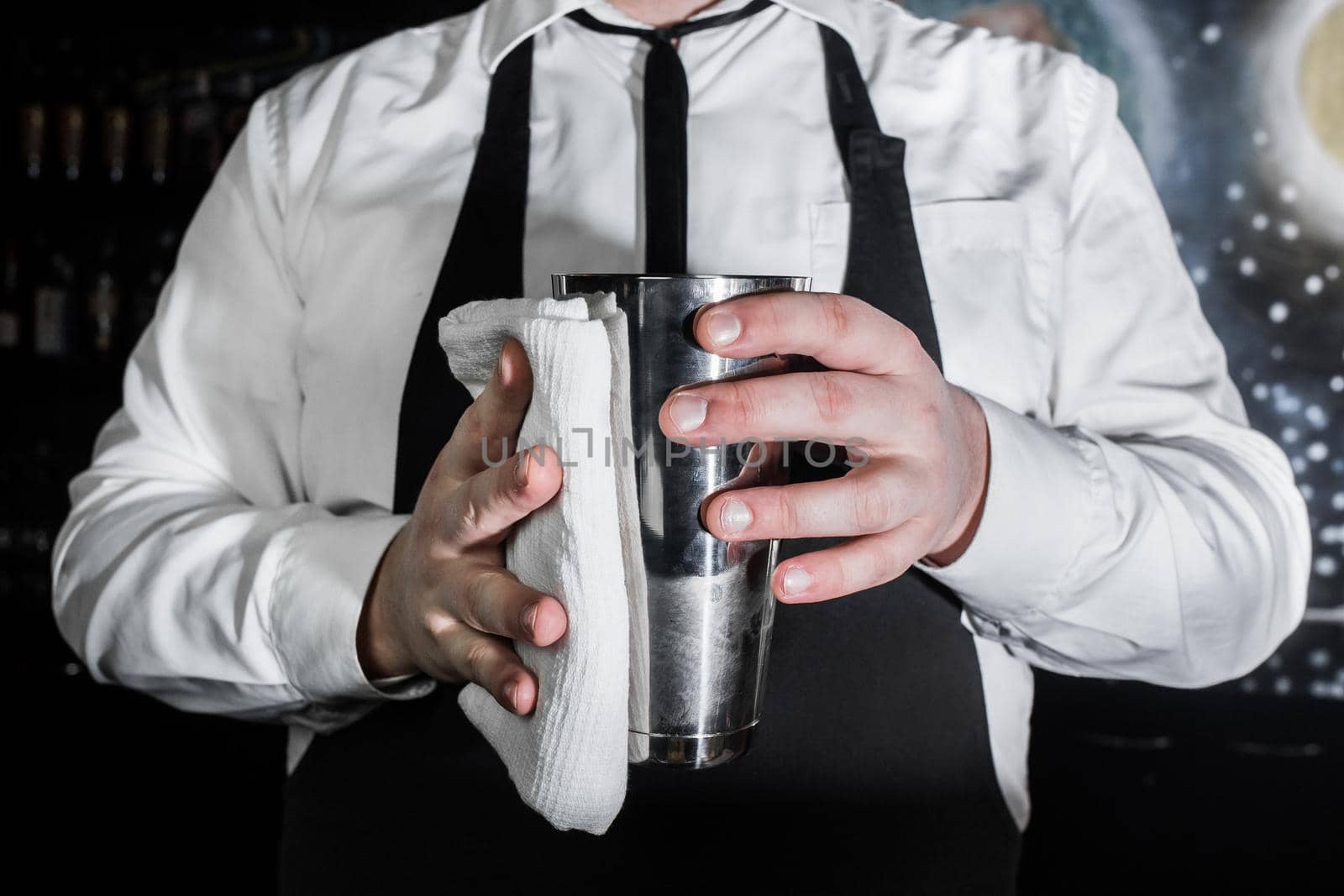 A professional bartender holds a metal shaker tool for mixing and making alcoholic cocktails and wipes it with a white cloth.