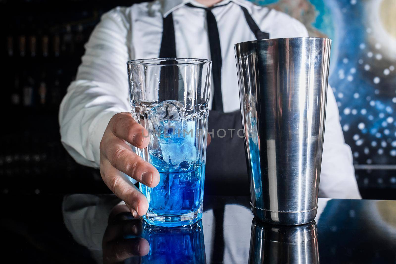 The hand of professional bartender takes a glass with ice and blue syrup stands on the bar counter, next to a tool for mixing and preparing alcoholic cocktails with a metal shaker.