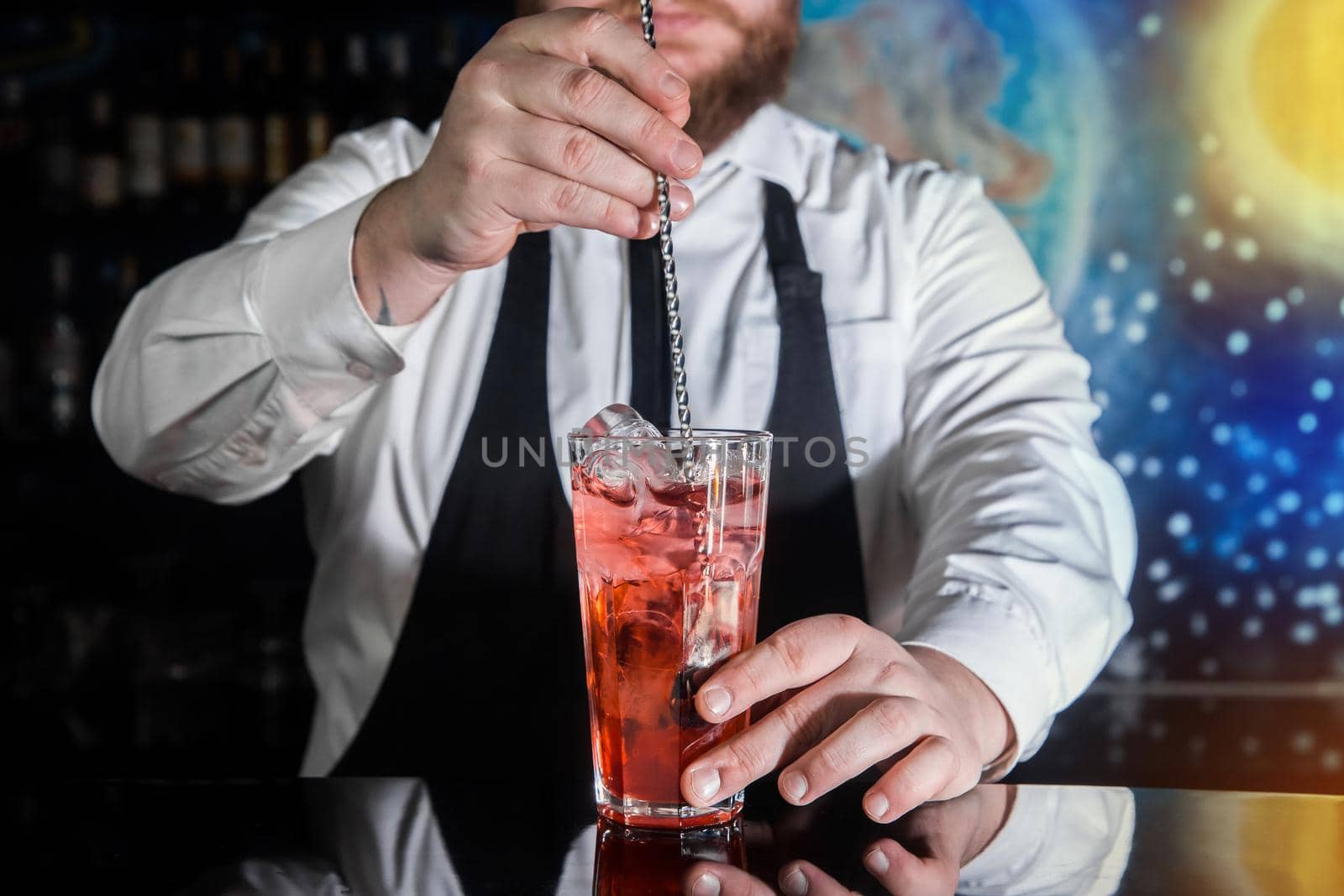 The hand of a professional bartender stirs the red syrup in an alcoholic cocktail with a bar spoon on the bar counter. The process of preparing an alcoholic beverage.