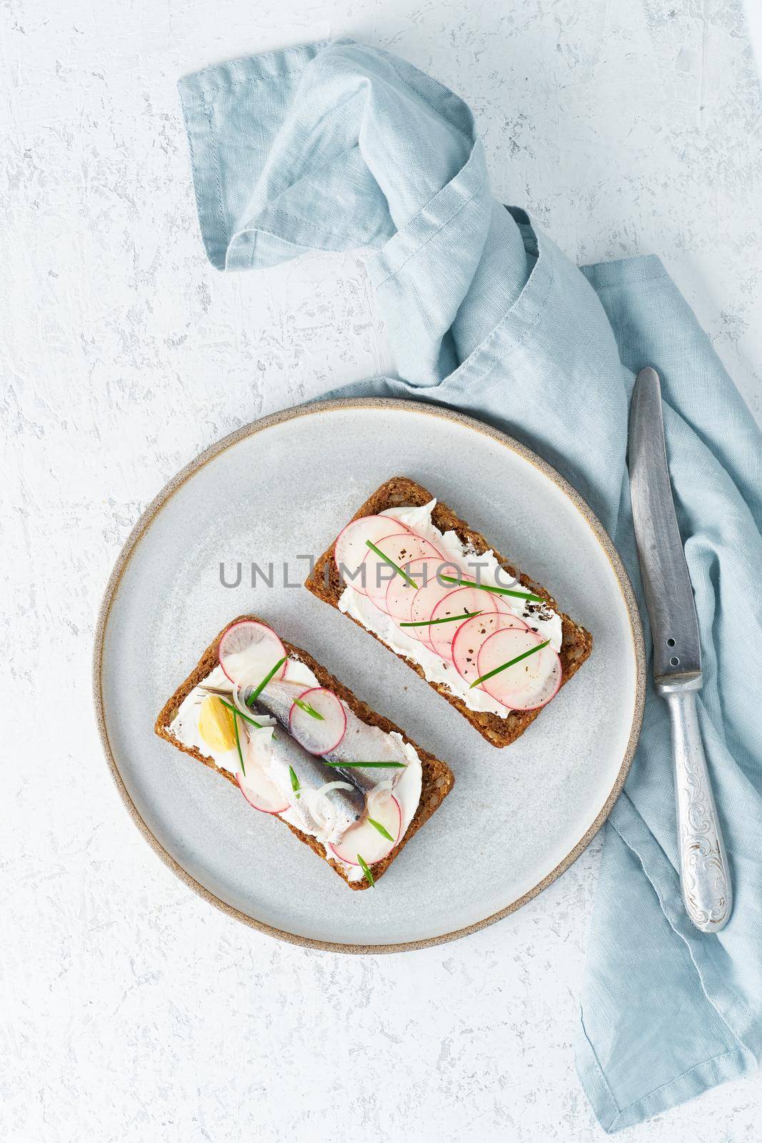 Savory smorrebrod, two traditional Danish sandwiches. Black rye bread with anchovy, radish, eggs, cream cheese on a grey plate on white stone table, vertical, top view