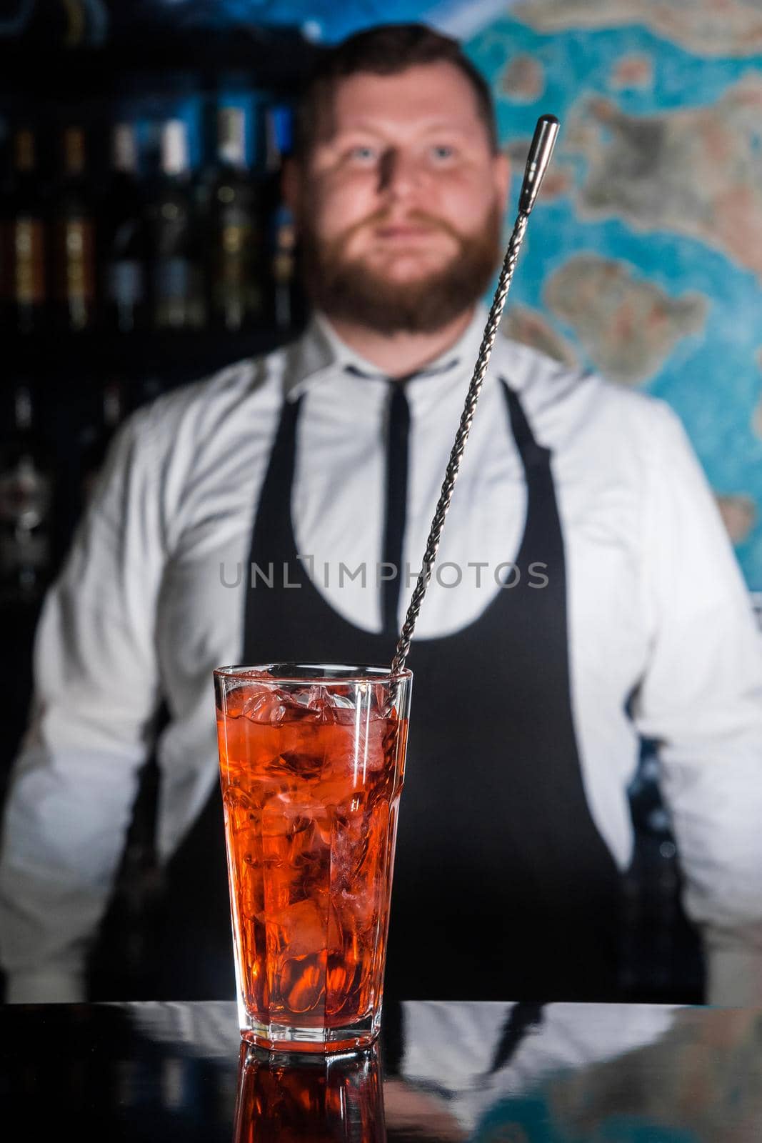 Red alcoholic beverage cocktail in glass with a bar spoon in front of a bearded bartender at the bar counter.