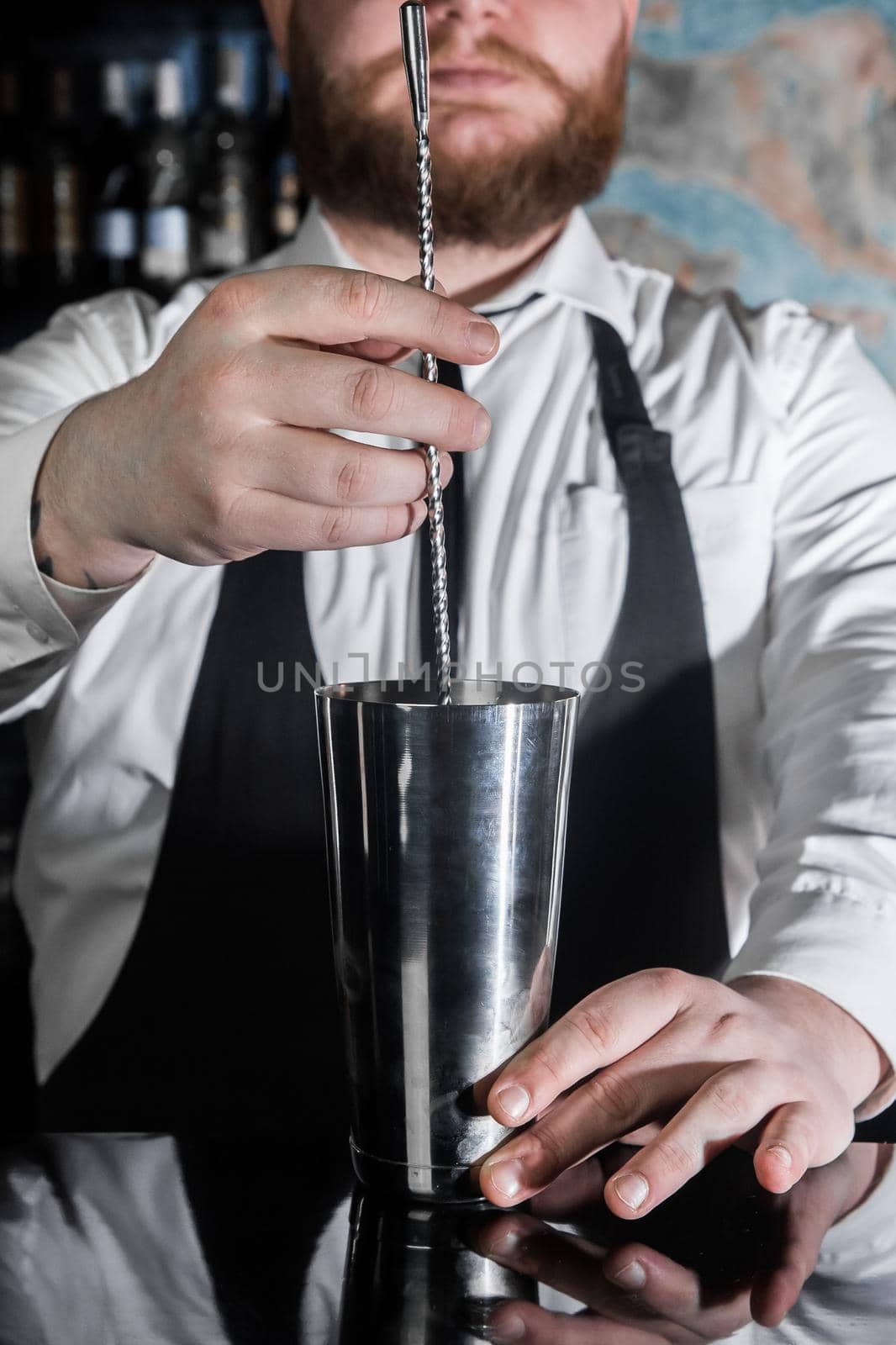 The hands of a professional bearded bartender interfere with a bar spoon the contents of a metal shaker tool for preparing and stirring alcoholic cocktails.