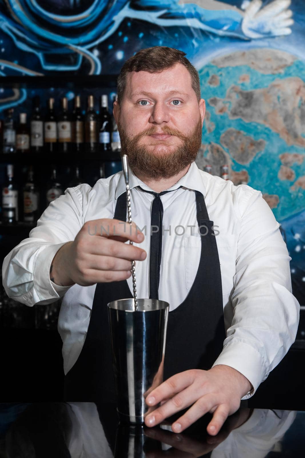 The hands of a professional bearded, adult bartender interfere with a bar spoon the contents of a metal shaker tool for preparing and stirring alcoholic cocktails by AYDO8