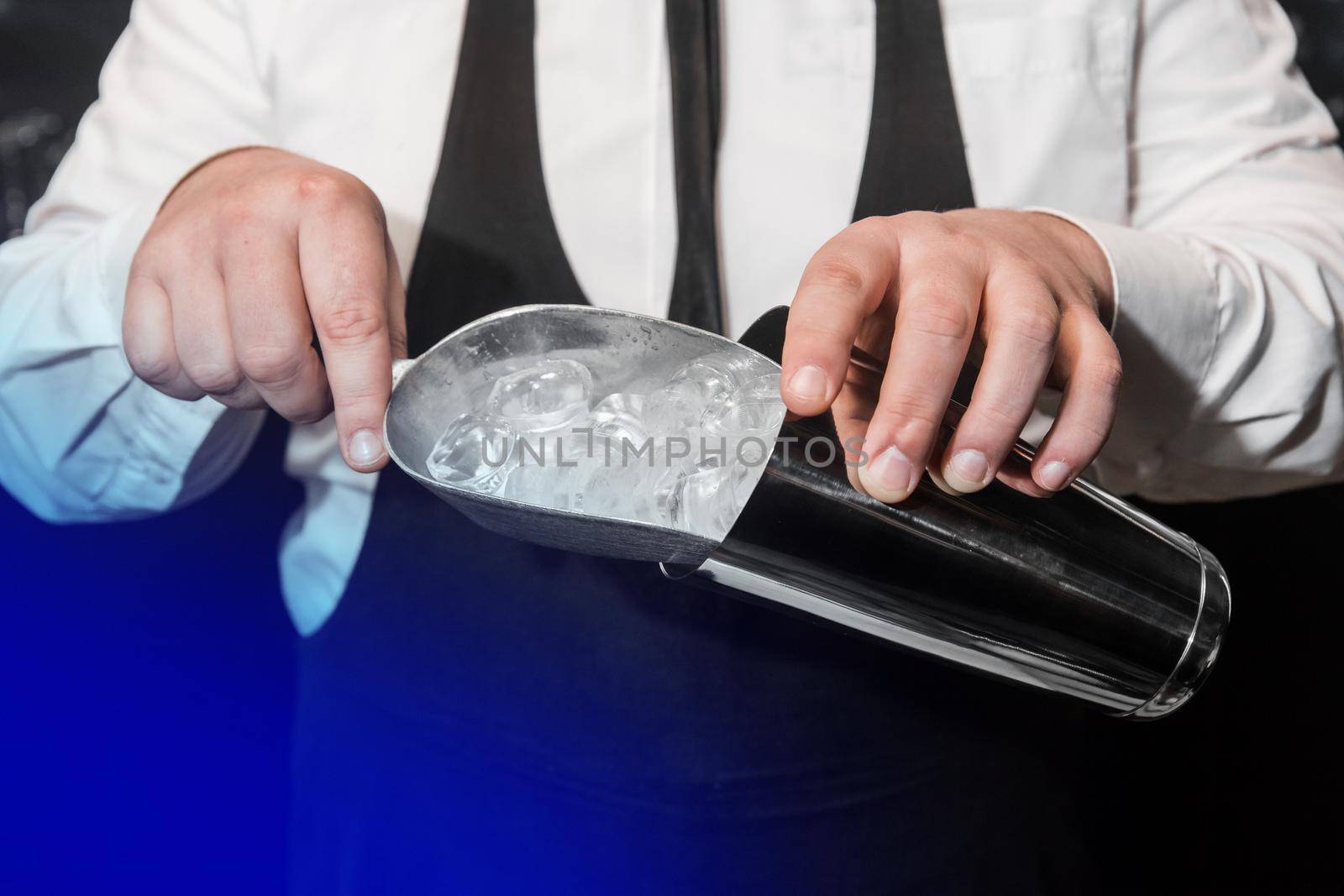 The hands of a professional bartender add ice in a scaple to a tool for preparing and mixing alcoholic cocktails, a metal shaker.