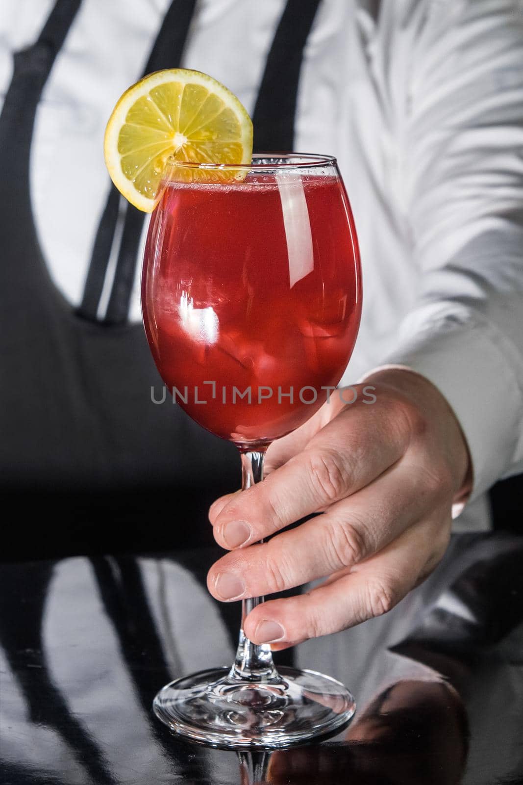 Professional bartender hand serves red alcoholic beverage in glass cocktail decorated with a slice of lemon on the bar counter.