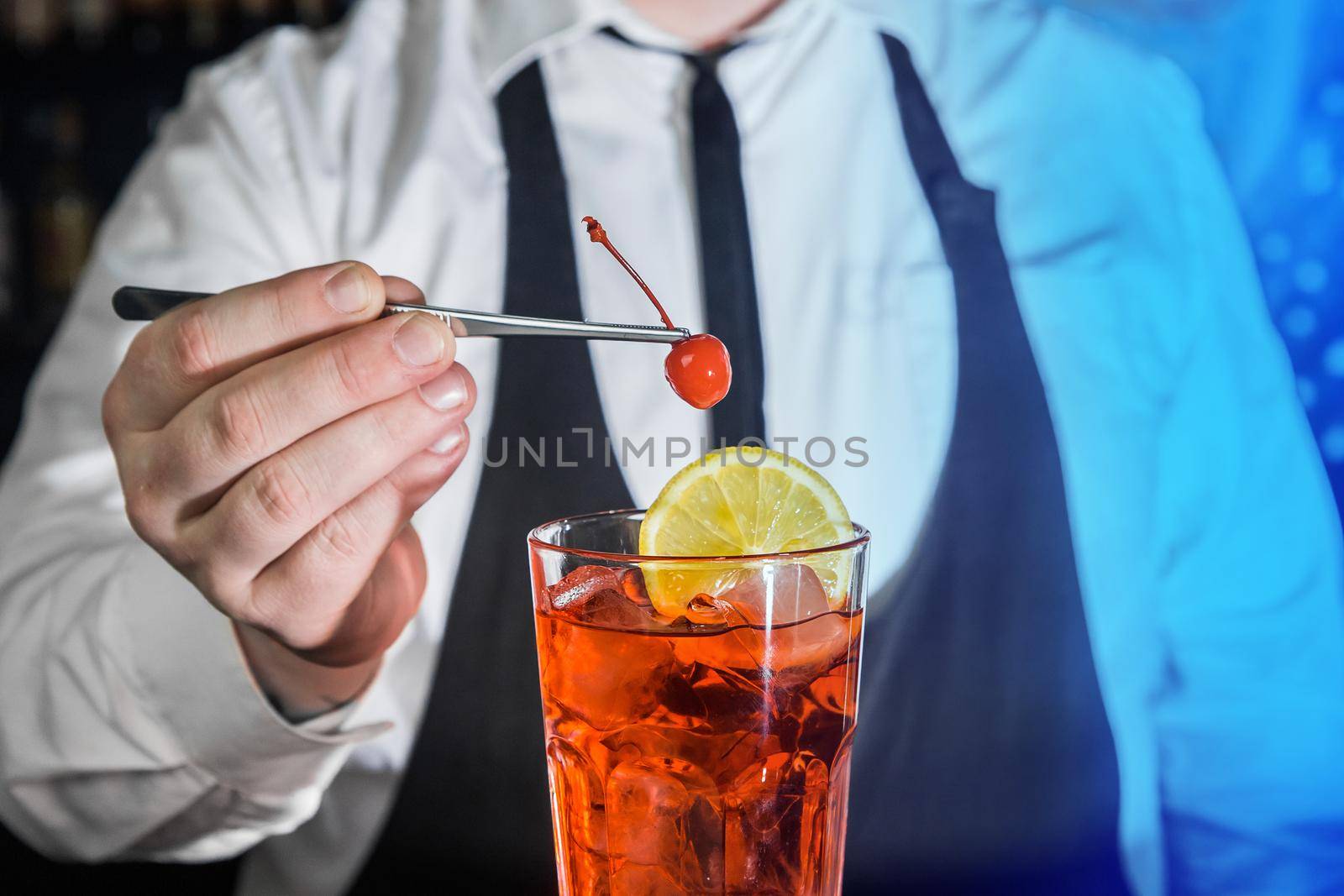 A professional bartender puts a cherry in a red chilled alcoholic cocktail with bar tweezers in glass at a nightclub counter.