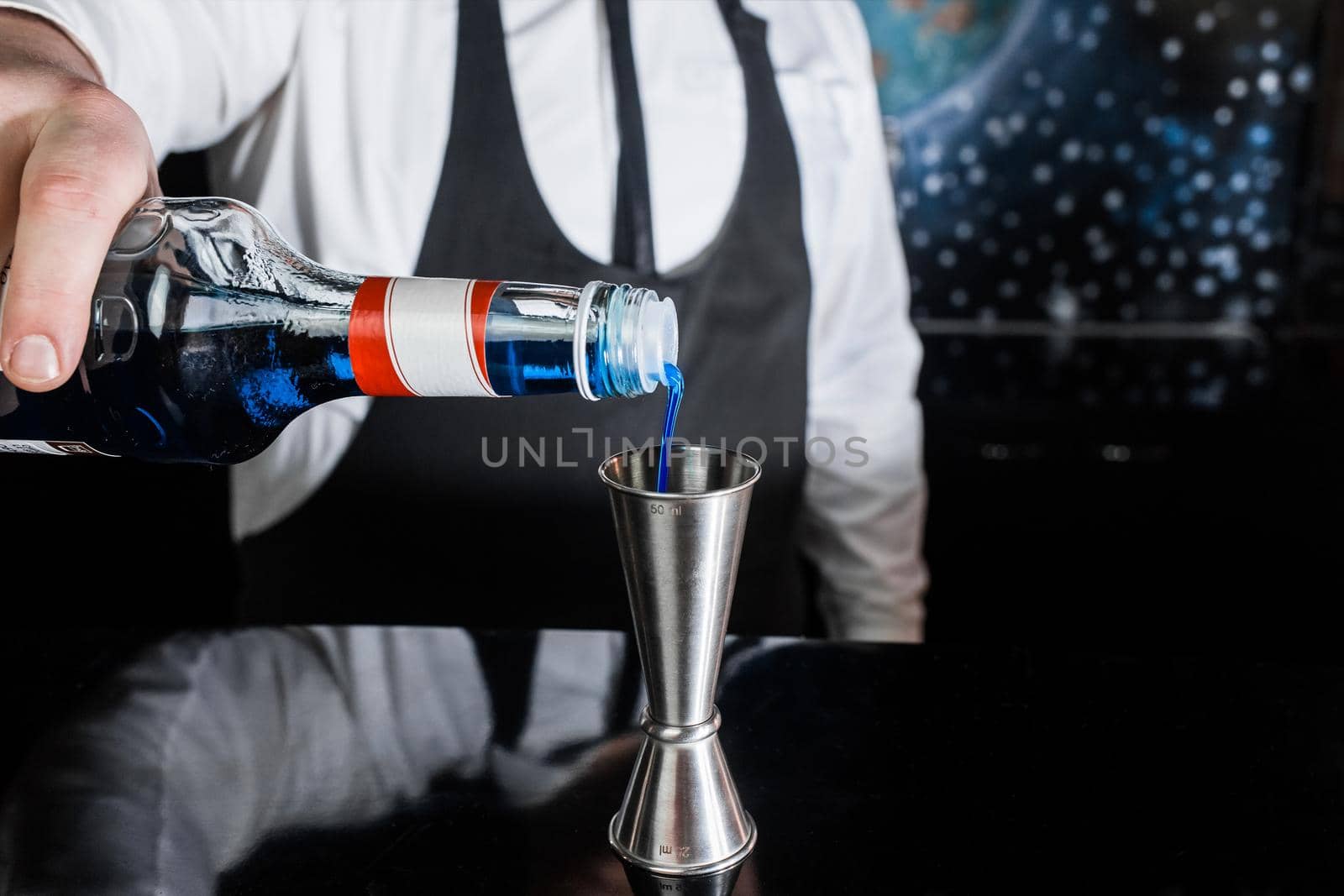 The hand of a professional bartender pours blue syrup into a tool to control the ingredients added to a jigger cocktail or measuring cup. The process of preparing a cocktail.