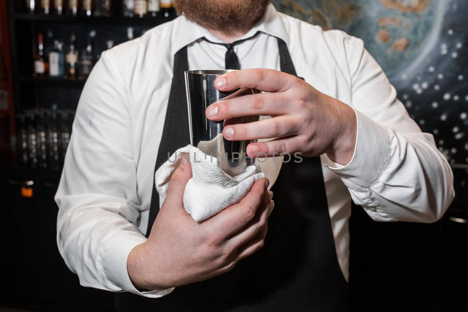 A professional bartender holds a metal shaker tool for mixing and making alcoholic cocktails and wipes it with a white cloth.