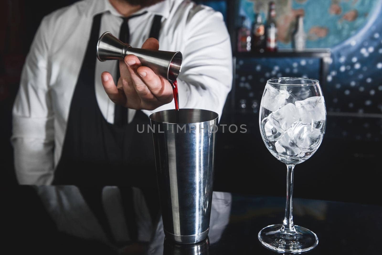 The hands of a professional bartender pour syrup into a measuring glass of jigger in a metal tool for preparing and stirring alcoholic cocktails of shaker drinks, and glasses with ice cubes.