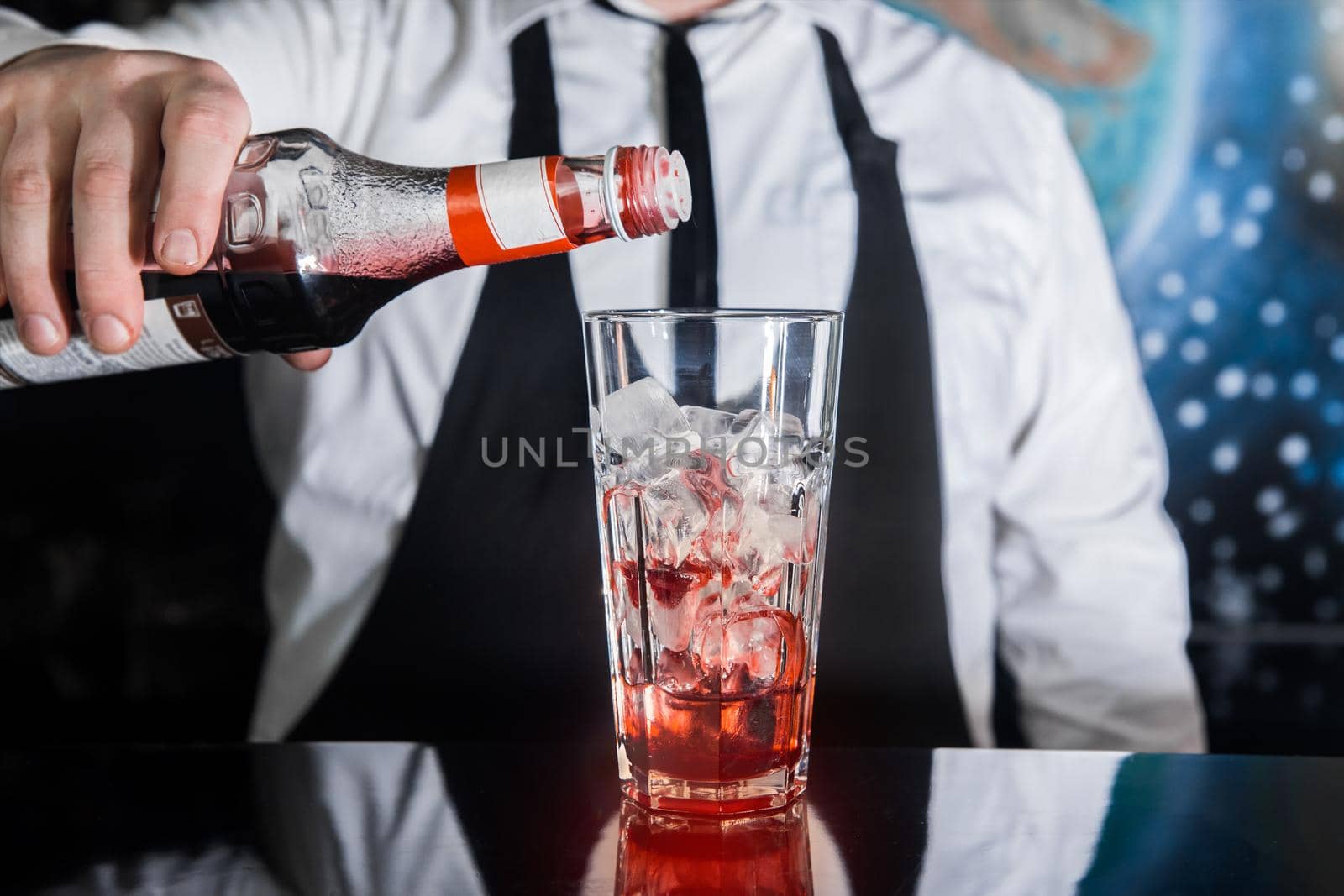 The hand of a professional bartender pours red syrup into a glass of ice cubes. The process of preparing an alcoholic cocktail.