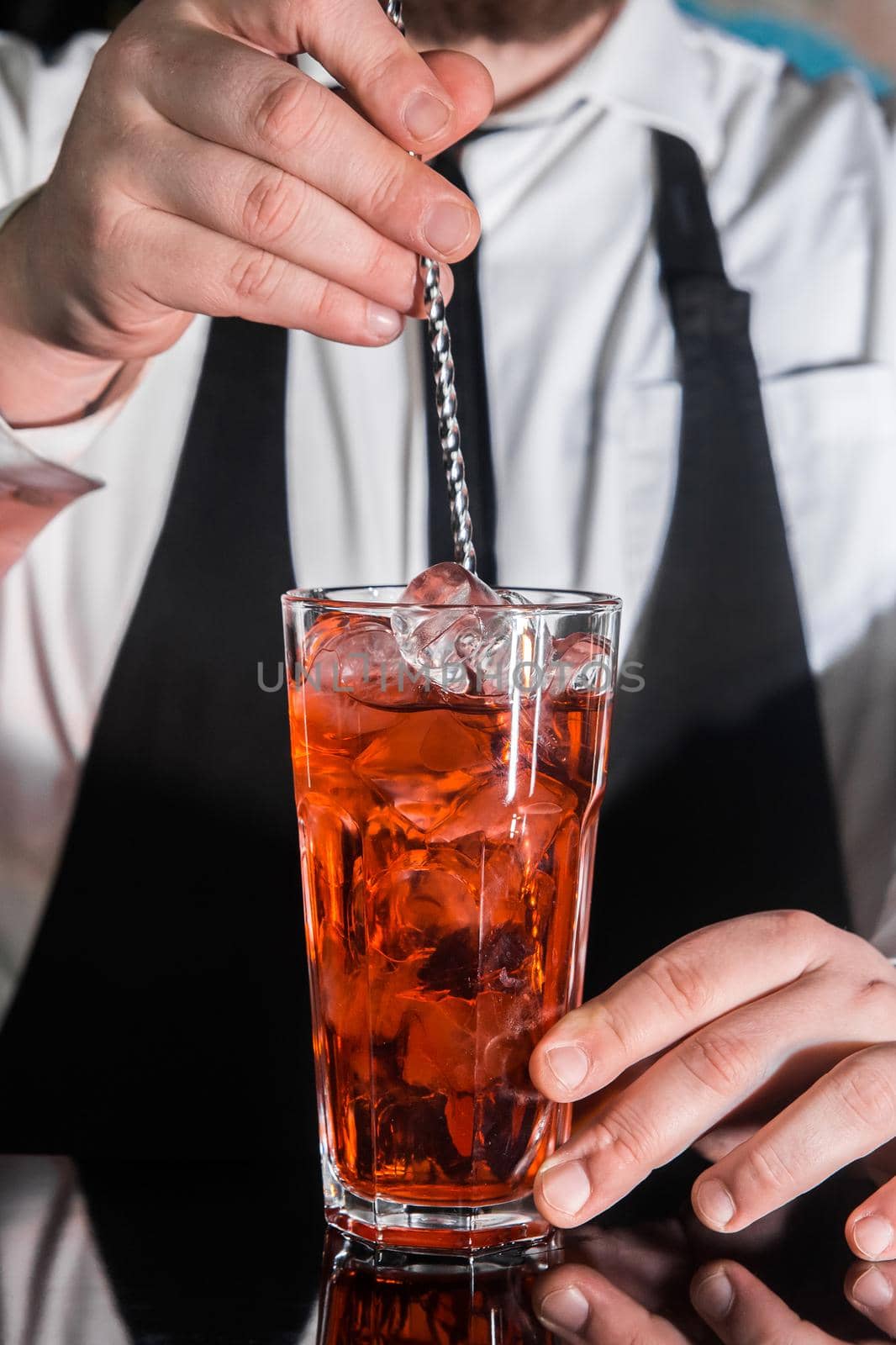 The hand of a professional bartender stirs the red syrup in an alcoholic cocktail with a bar spoon on the bar counter. The process of preparing an alcoholic beverage by AYDO8