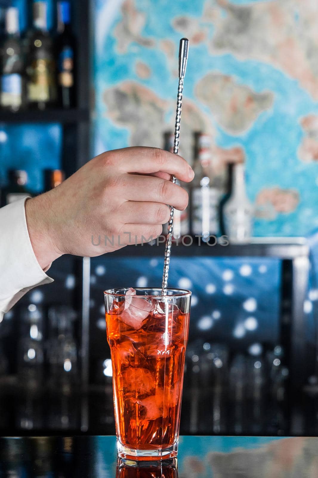 The hand of a professional bartender interferes with a bar spoon red alcoholic cocktail chilled drink in glass on the bar counter, close-up.