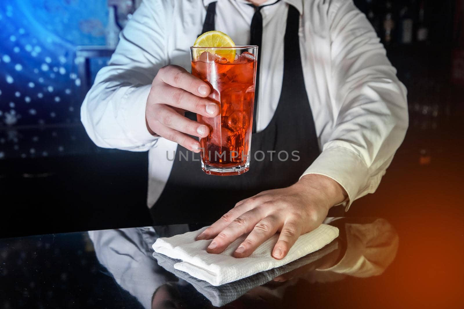 A professional bartender holds in his hand a red alcoholic cocktail, chilled drink in glass decorated with a slice of sliced lemon and wipes the bar counter with white cloth for serving by AYDO8