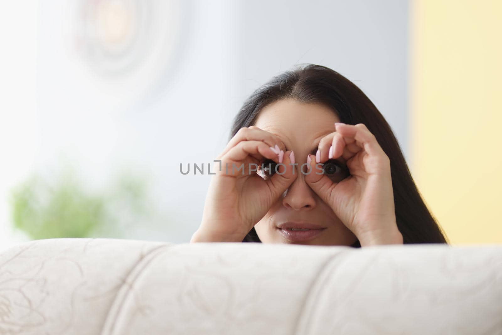 Portrait of cheerful woman peeking from behind sofa, female hands make binoculars over eyes. Adult fooling around, have fun. Game, entertainment concept