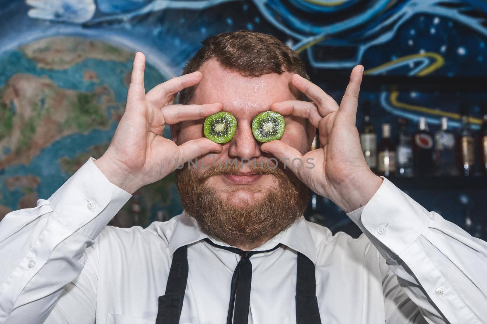 Adult funny bearded positive man professional bartender holds halves of sliced kiwi in his hands at the face.