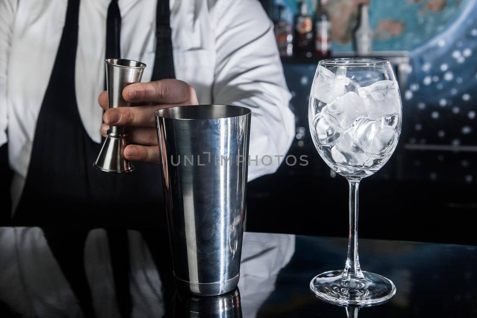 The hands of a professional bartender pour syrup into a measuring glass of jigger in a metal tool for preparing and stirring alcoholic cocktails of shaker drinks, and glasses with ice cubes.