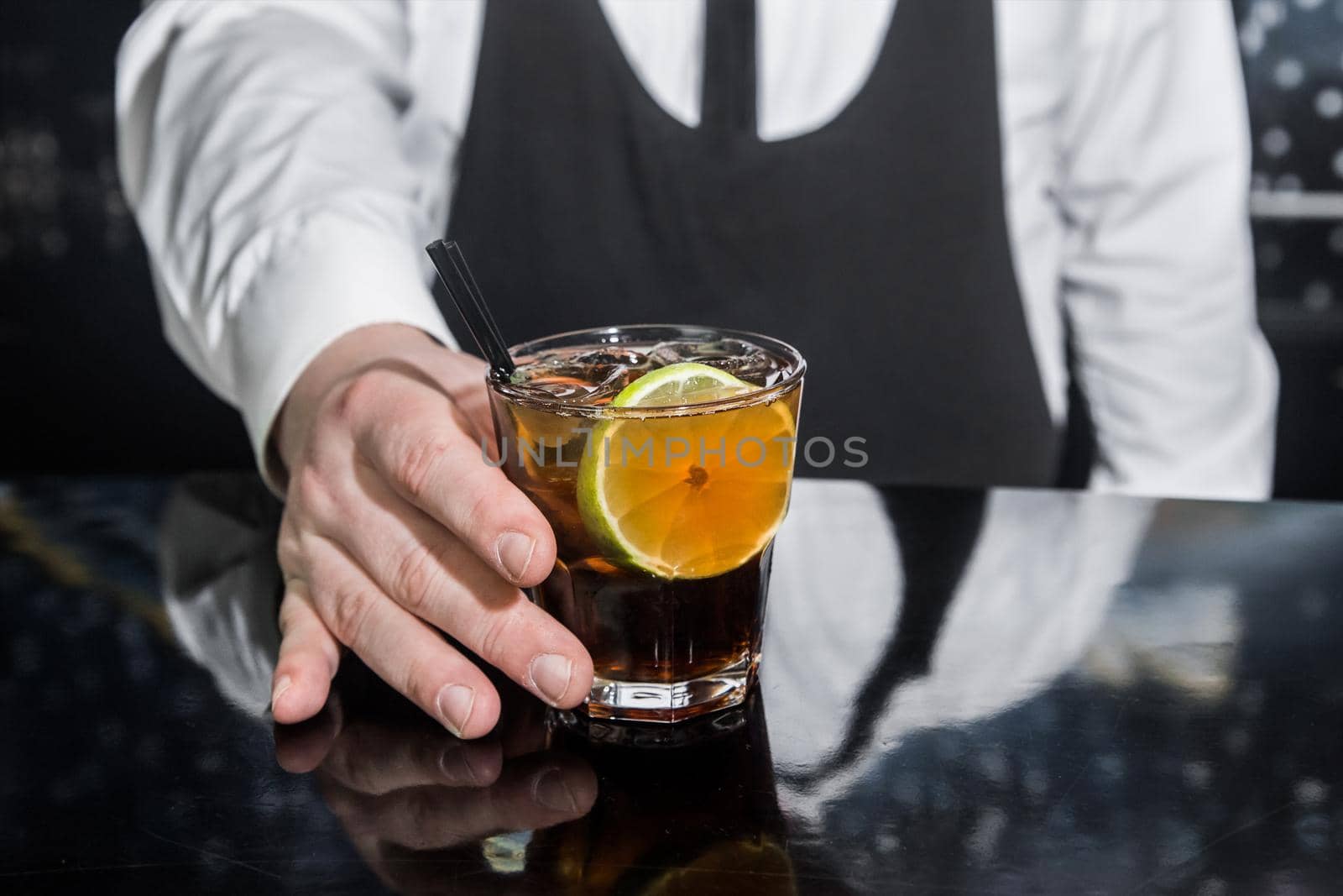 The hand of a professional bartender serves an alcoholic beverage cocktail whiskey with cola and a piece of lime on the bar counter, close-up.