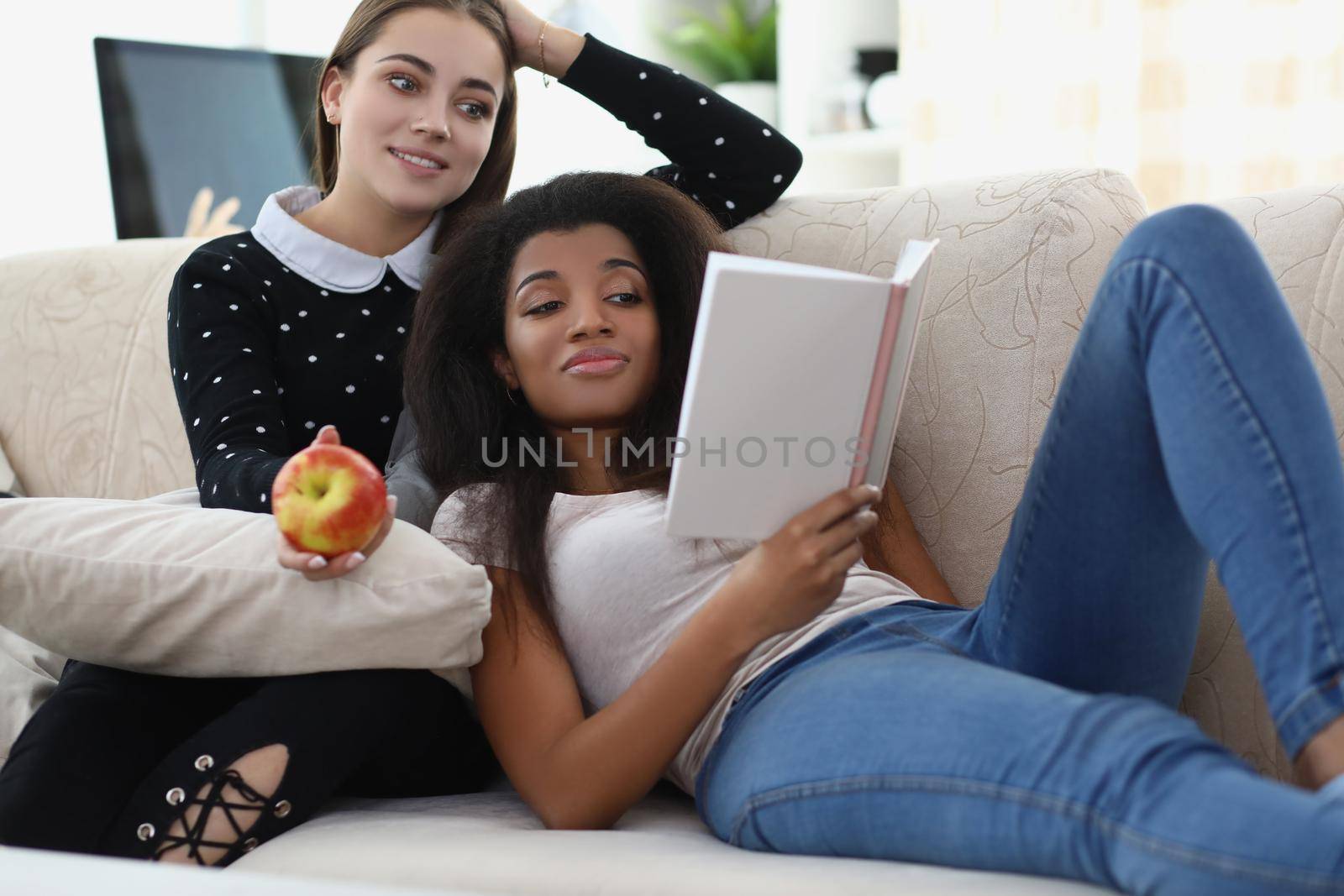 Portrait of best friends reading book chilling on sofa, apple for snack, relaxing atmosphere. Expand knowledge in literature. Leisure, friendship concept