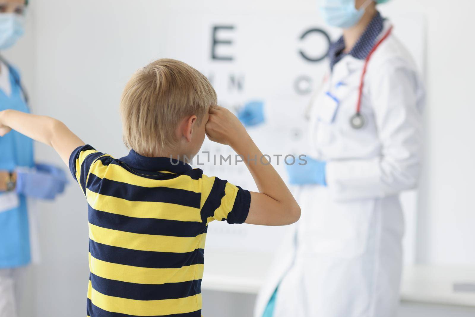 Portrait of boy on appointment in clinic, oculist cabinet, sight diagnostic closing eye and say out loud letter on board. Health, ophthalmology concept