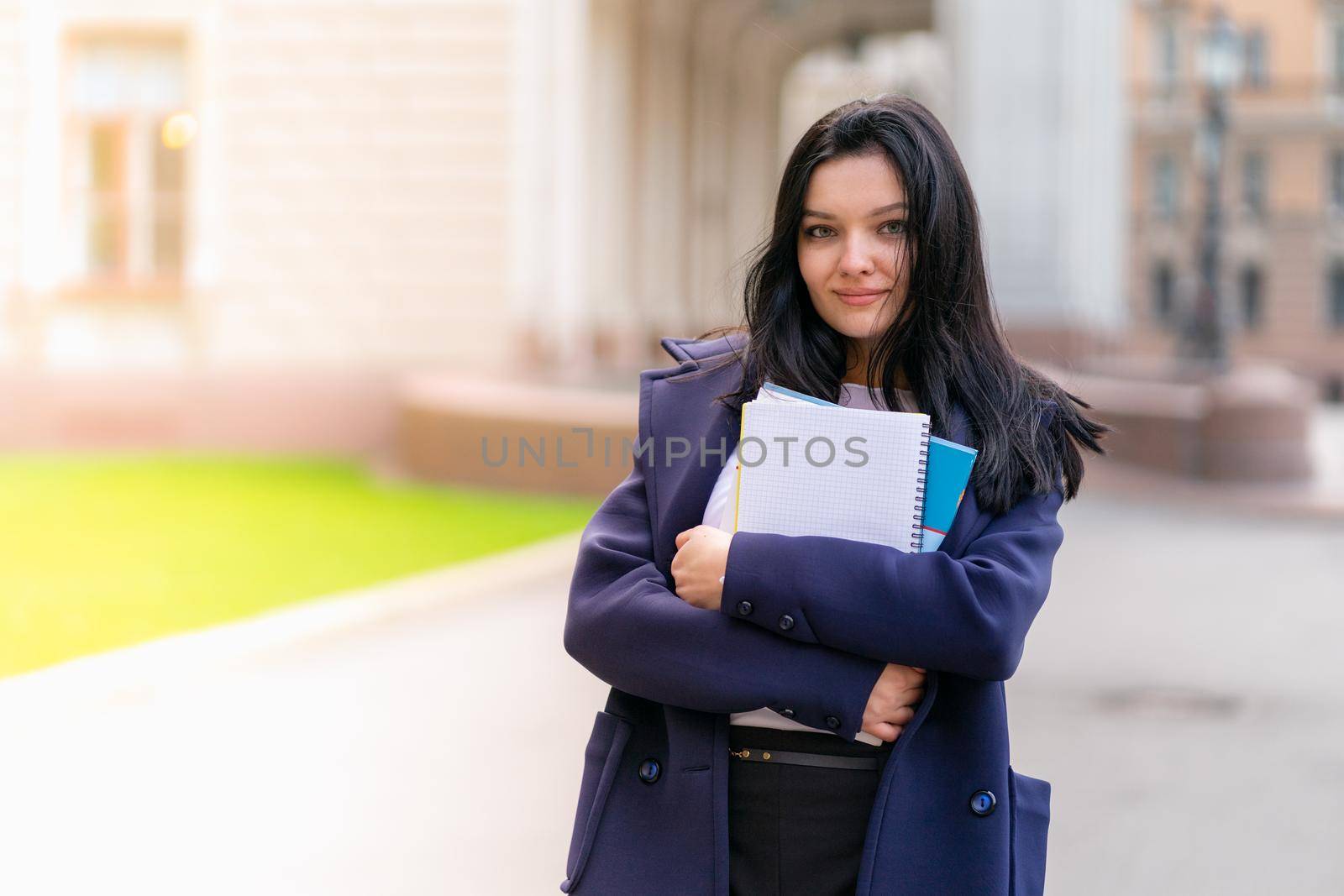 Beautiful smiling girl brunette student holding notebooks and textbooks, stands at University on street of St. Petersburg. A charming woman with long dark hair is studying at a course or at College