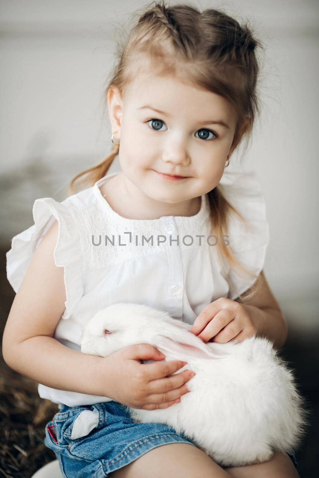 Portrait of little cute kid with braids on head looking at camera. Adorable small girl in casual clothes holding her fluffy sleepy pet on khees. Happy pretty child touching white rabbit and playing.