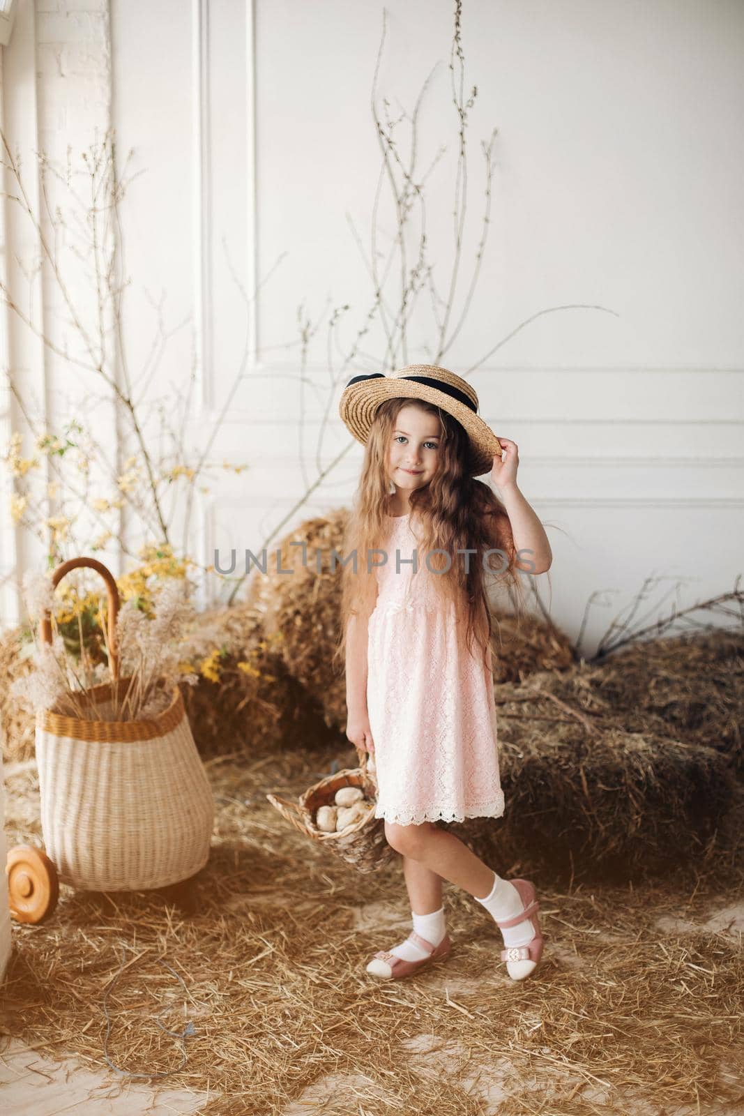 Front view of beautiful girl wearing pink dress and hay hat keeping basket with little chick. Cute female child looking aside and smiling while walking in garden in village. Concept of childhood.