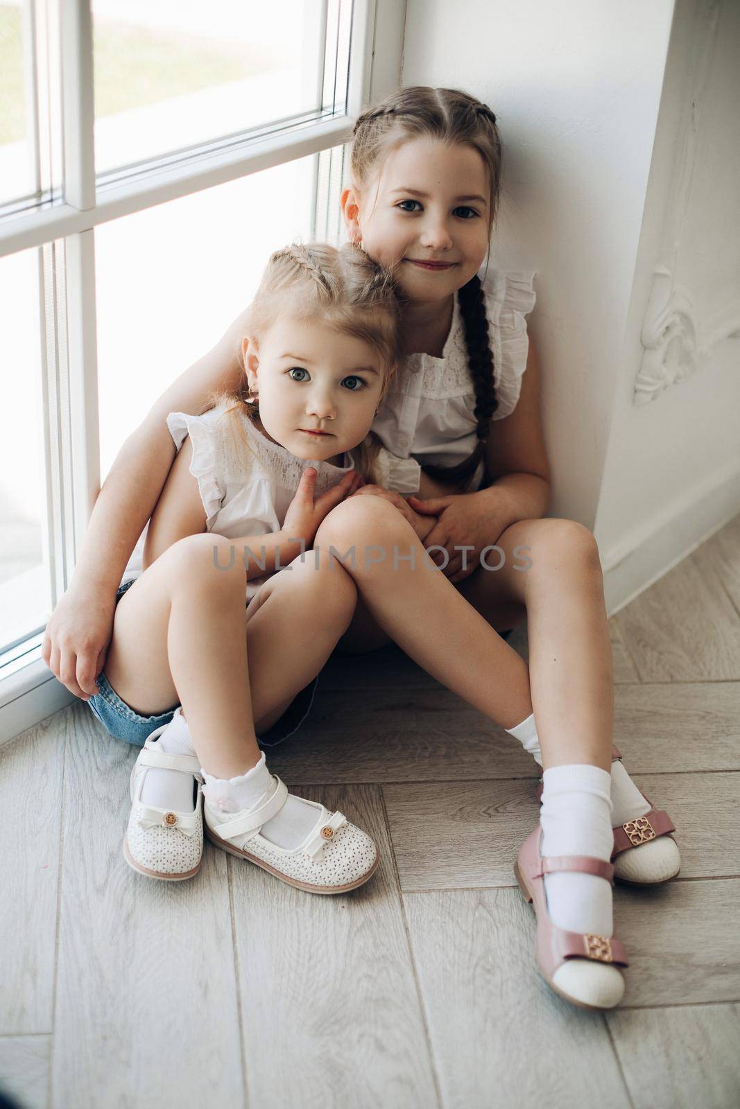 View from above of two beautiful sisters sitting together near window and hugging each other. Cute girls looking at camera, smiling and posing at home. Concept of childhood and family.