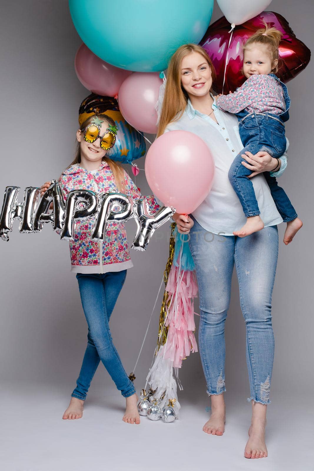 Front view of happy family looking at camera and posing while celebrating birthday party. Young mother keeping little daughter while girl standing near and keeping colorful balloons. Concept of fun.