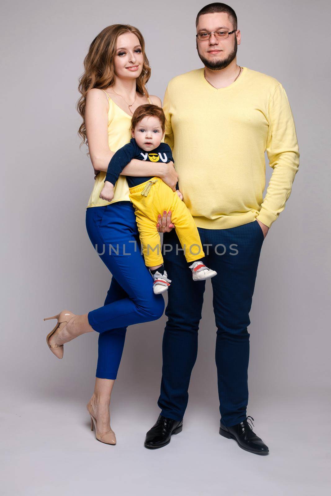 Portrait of happy family from three people wearing same coloured clothes. Blonde beautiful mother smiling and holding on hands surprised child. Handsome father in glasses hugging lovely wife and son.