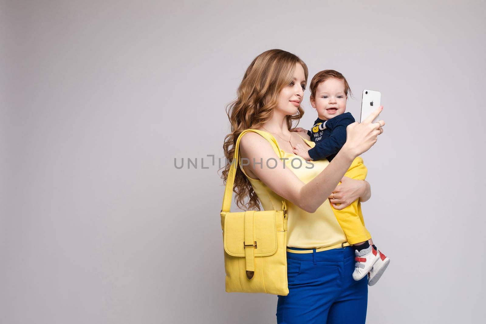 Side view of pretty mother walking with little kid and taking selfie. Young woman keeping child on hands and posing on isolated background in studio. Concept of childhood and family outfit.