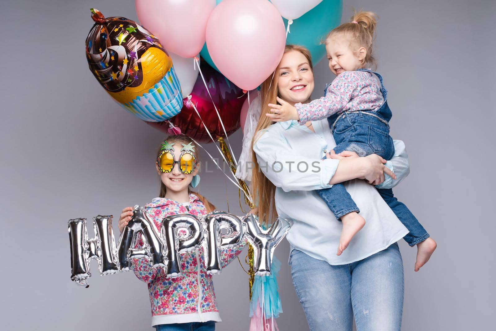 Front view of happy family looking at camera and posing while celebrating birthday party. Young mother keeping little daughter while girl standing near and keeping colorful balloons. Concept of fun.