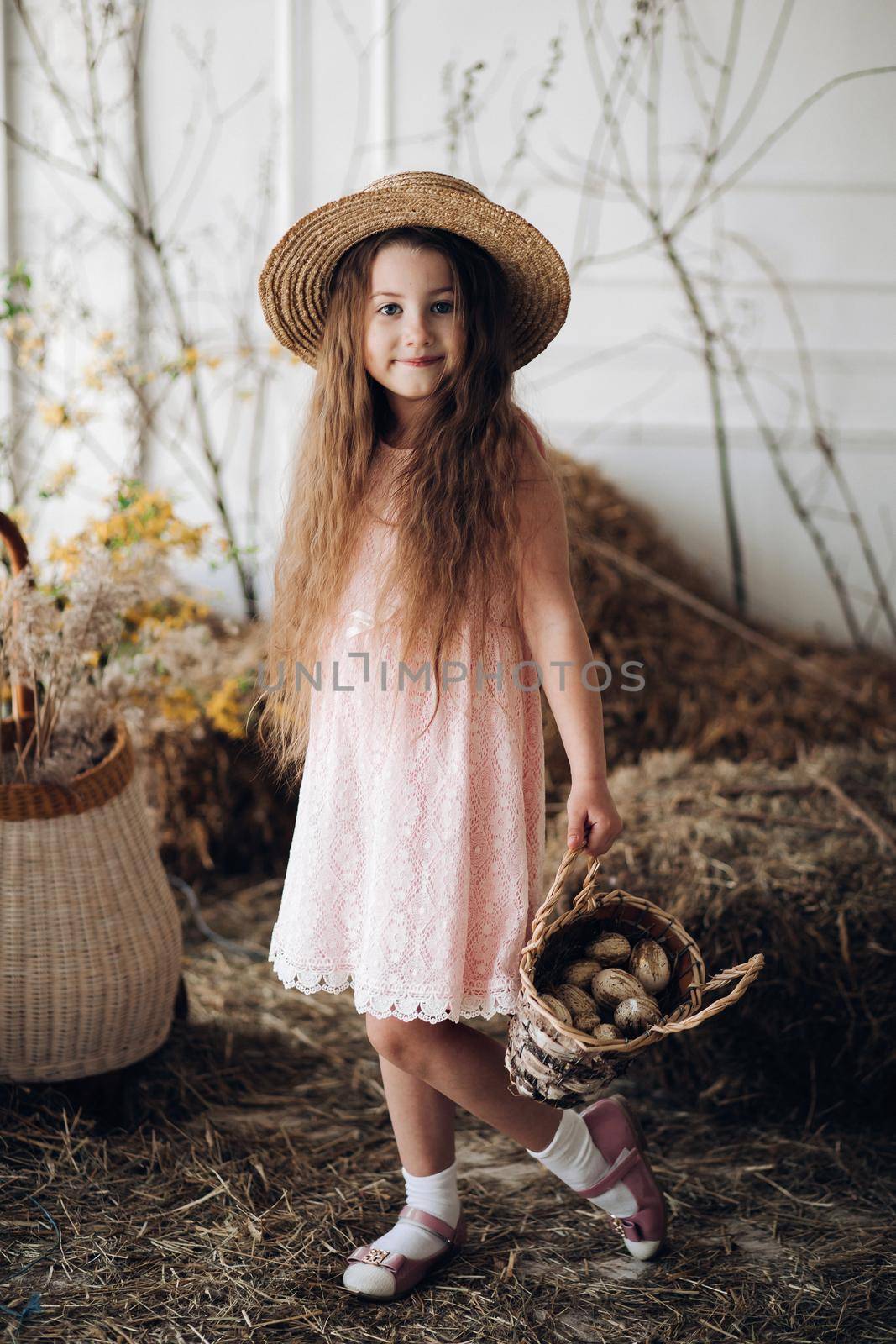 Portrait of little charming kid in peachy elegant dress and hat shyly posing at camera. Small cute girl standing among decoration and bunches of hay. Sweet lovely child with long hair holding hand up.