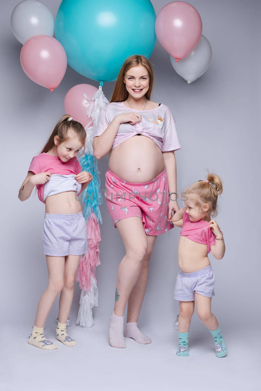 Stunning pregnant woman showing her belly. Her two daughters looking at their tummy too. They all wearing pyjamas. Studio portrait of mother and two daughters demonstrating bellies.