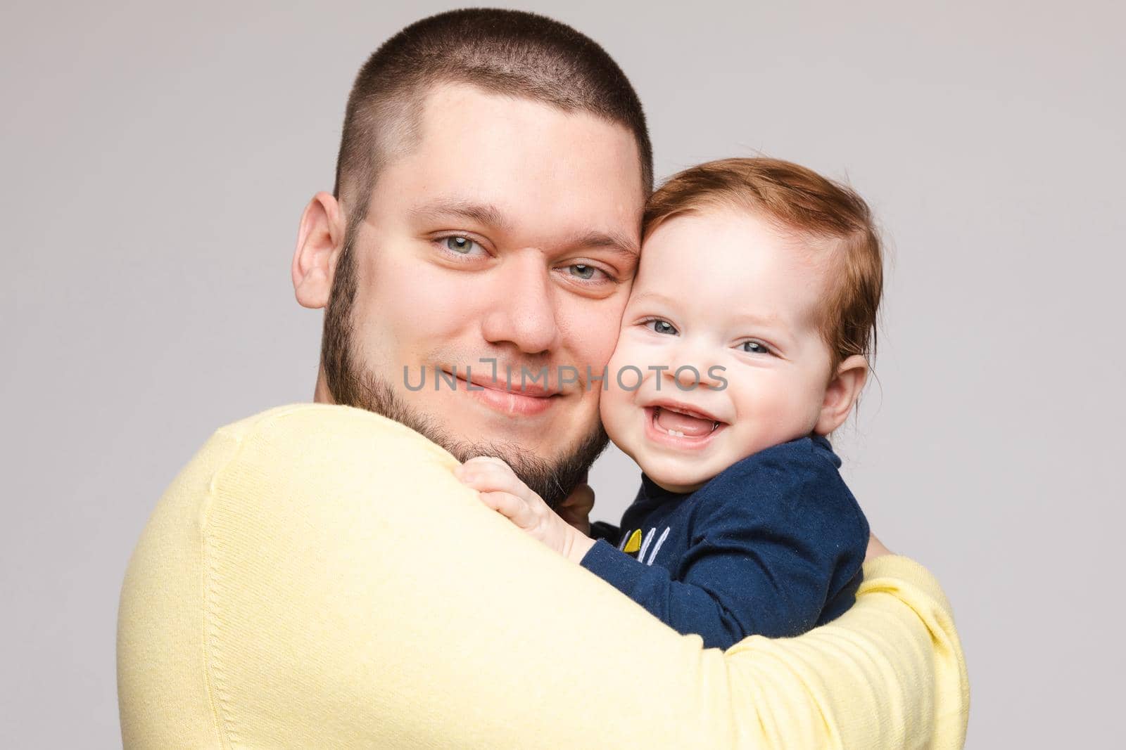 Portrait of happy father in yellow sweater posing with his lovely smiling child. Athletic and young men with beard holding red haired little boy. Handsome brunette dad hugging sweet and cute son.