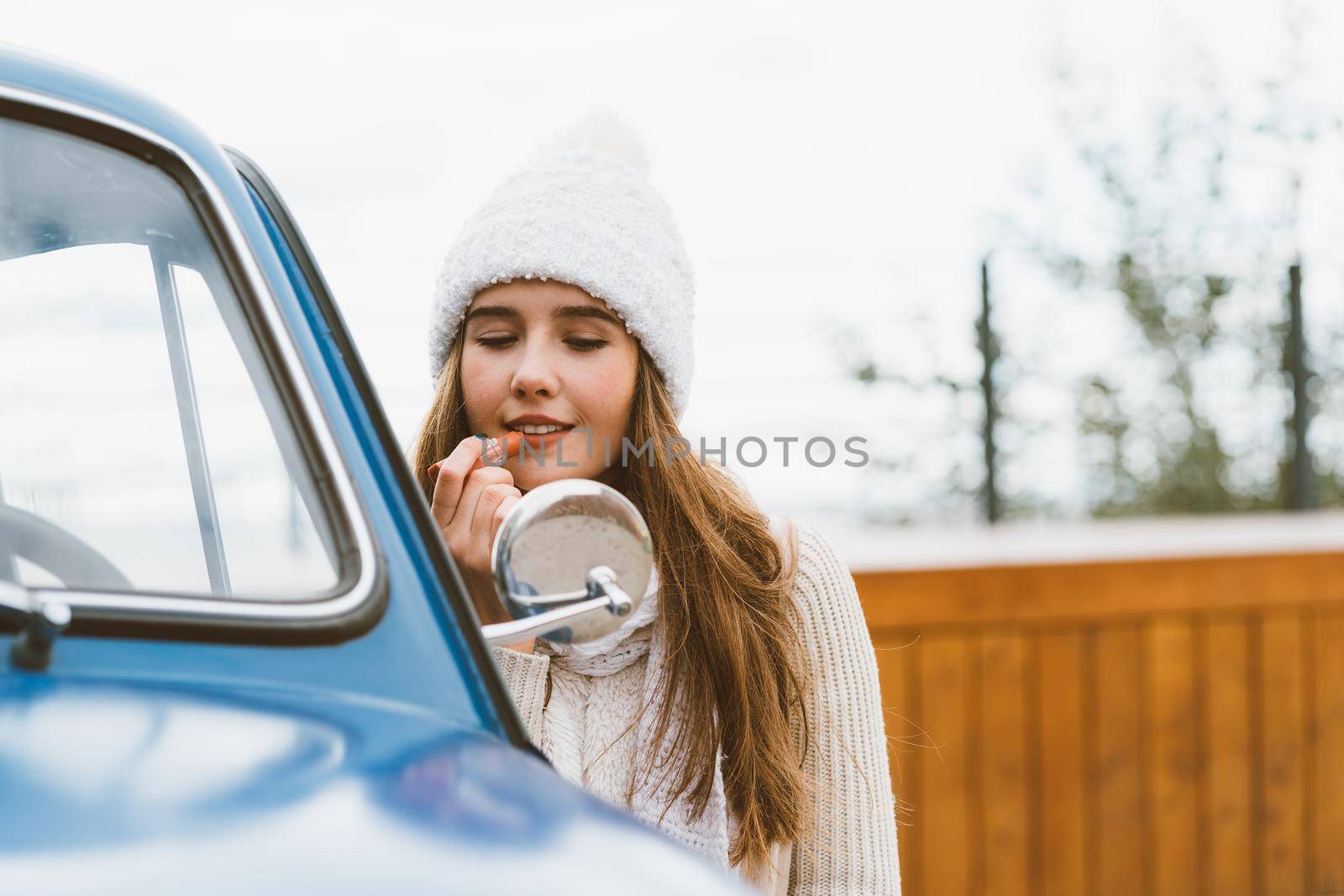 A beautiful young girl looks in the car mirror and paints her lips. Woman with long dark thick hair next to blue retro car in autumn or winter, copy space by NataBene
