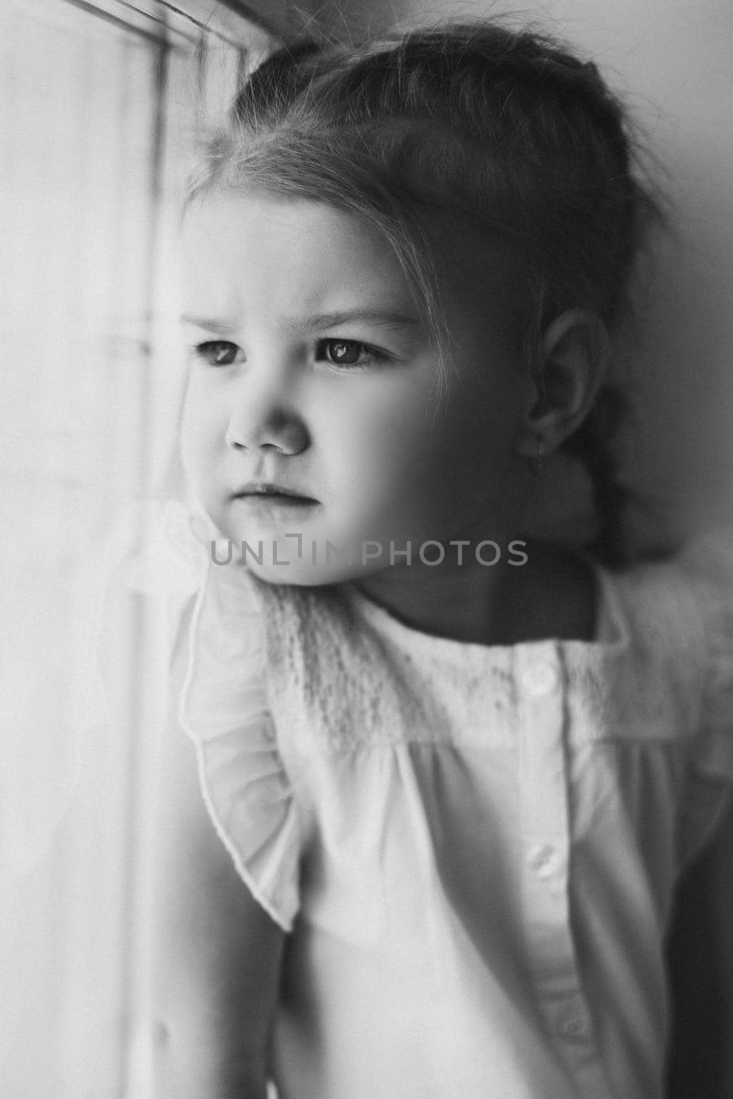 Portrait of little cute kid leaning on wall and looking at camera. Small pretty girl standing near big window and smiling. Adorable child with braid in casual clothes holding arms crossed on chest.