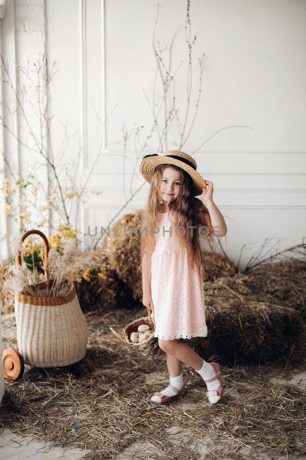 Portrait of little charming kid in peachy elegant dress and hat shyly posing at camera. Small cute girl standing among decoration and bunches of hay. Sweet lovely child with long hair holding hand up.