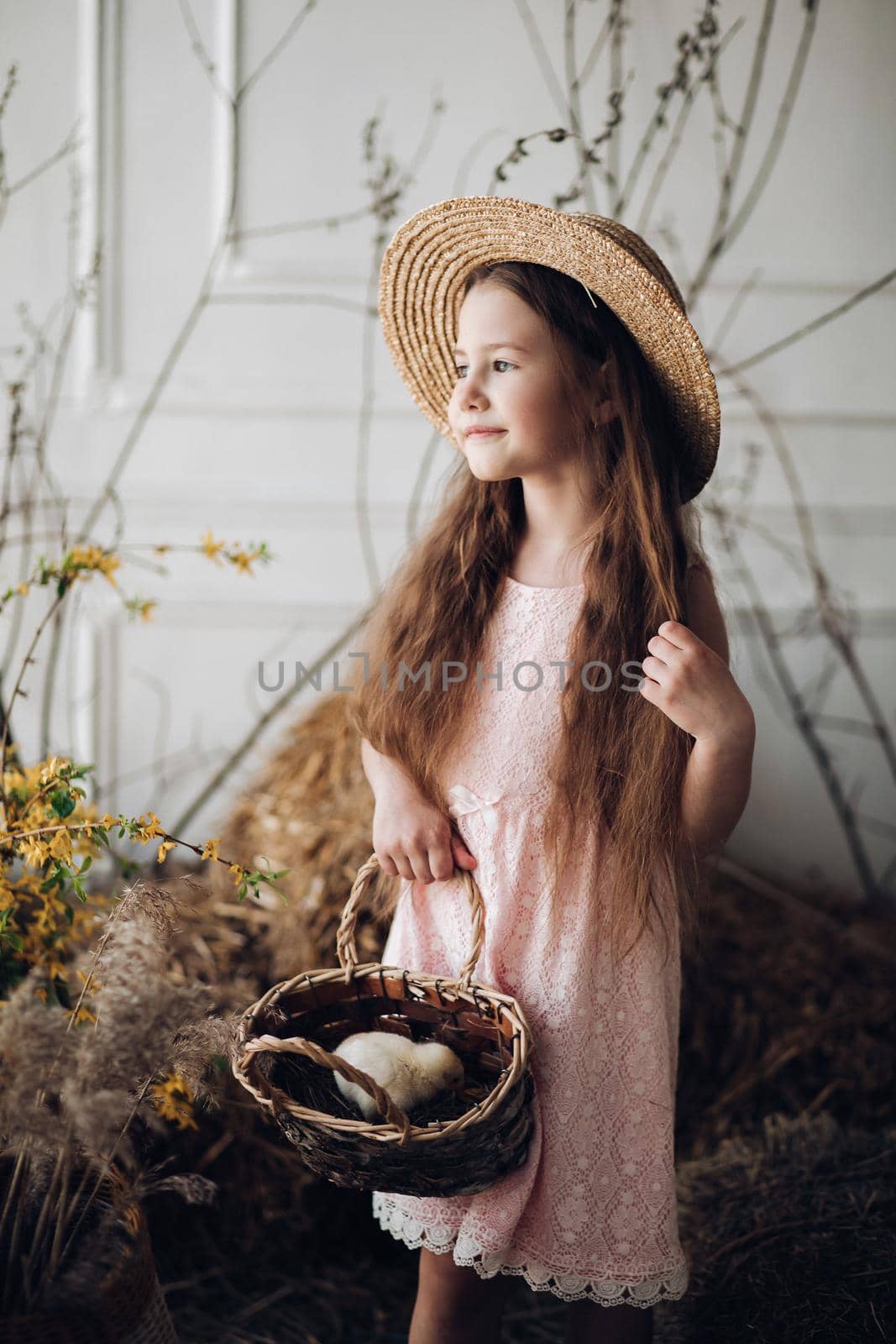 Front view of beautiful girl wearing pink dress and hay hat keeping basket with little chick. Cute female child looking aside and smiling while walking in garden in village. Concept of childhood.